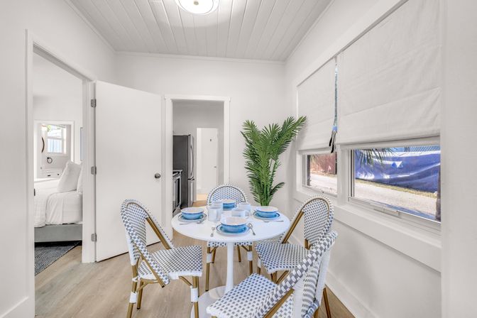 Bright coastal-style dining nook with round white table set with blue dishes, woven bistro chairs, potted palm, hardwood floors, and sunlit windows with roman shades