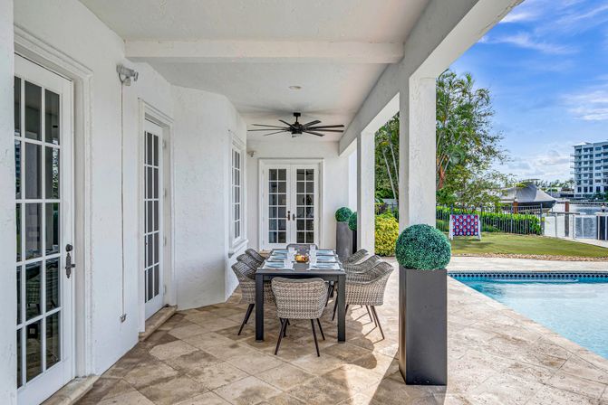 Covered waterfront patio with modern six-seat outdoor dining set on stone tile beside a swimming pool, ceiling fan, French doors and marina view for relaxed coastal poolside dining.