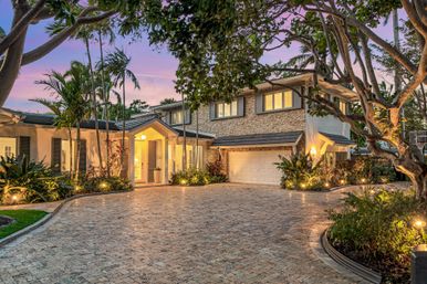 Luxury two-story home at dusk with stone facade, illuminated entryway and double garage, circular paver driveway, palm trees and lush tropical landscaping under a pink sunset sky.
