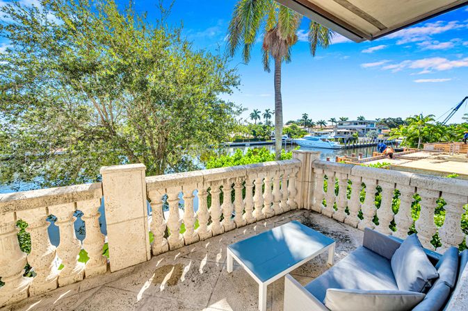 Sunny tropical waterfront balcony with stone balustrade, cozy outdoor sofa and coffee table overlooking a canal marina, palm trees and a docked yacht.