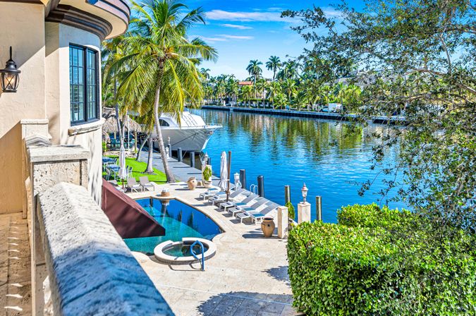 Sunny balcony view of a luxury waterfront patio with pool and spa, lounge chairs, palm trees and a yacht on a calm canal.