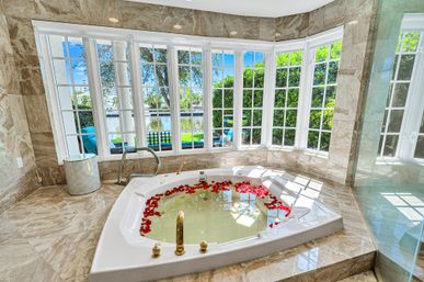 Sunlit luxury bathroom with marble tile and a sunken jacuzzi tub filled with water and red rose petals, framed by large bay windows overlooking a lush waterfront garden and canal — spa-like, romantic view.