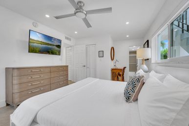 Bright airy modern bedroom with crisp white bedding and patterned throw pillows, wooden six-drawer dresser, wall-mounted landscape TV, ceiling fan, bedside lamp, small wooden desk and a large window with greenery outside
