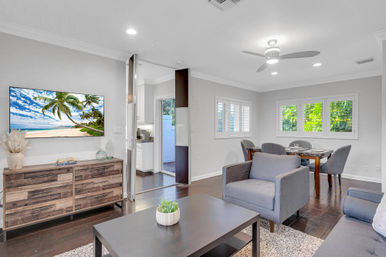 Bright modern open-plan living and dining room with dark hardwood floors, gray armchairs, wooden credenza, dining table by shuttered windows, and a wall-mounted TV showing a tropical beach for a vacation-vibe touch.