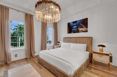 Sunlit luxury bedroom with plush king bed and velvet headboard, sparkling crystal chandelier, midcentury nightstands with golden lamps, beige curtains framing windows with palm-tree views, hardwood floors and bold blue artwork above the bed.