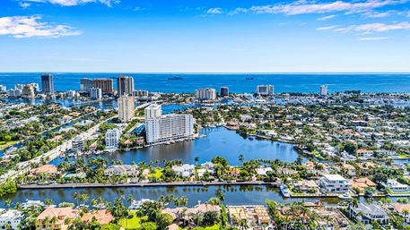 Aerial view of a sunny Florida coastal city with palm-lined canals, waterfront homes and marinas, mid-rise condos and the blue Atlantic Ocean on the horizon.