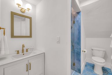 Chic compact bathroom with white walls and vanity, gold faucet and mirror trim, glass shower with blue tile, patterned blue floor tiles and white toilet