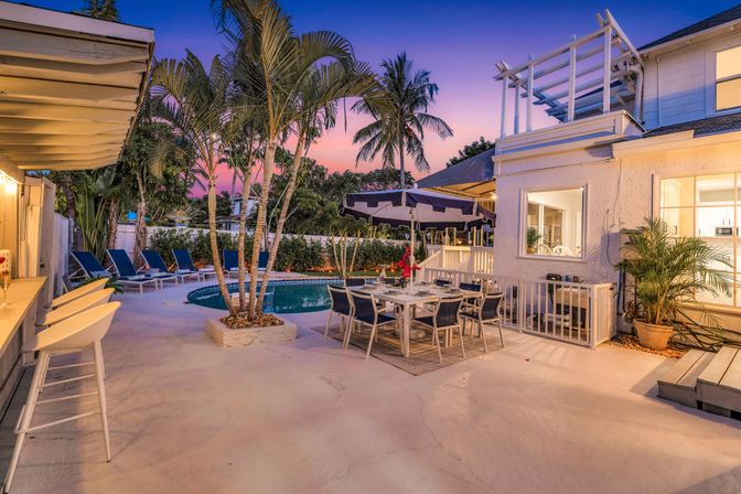 Tropical poolside patio at sunset with palm trees, lounge chairs, bar seating and an outdoor dining table beside a two-story home