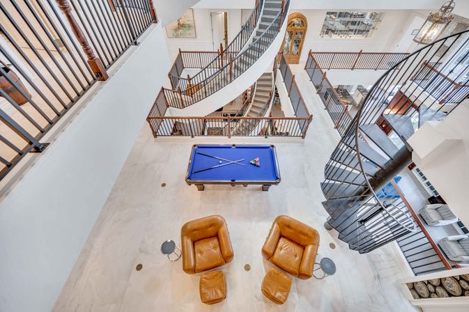 Aerial view of a modern multi-level open atrium with a black spiral staircase, wraparound balconies, blue pool table and two tan leather lounge chairs in a bright contemporary game room