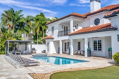 Sunlit Mediterranean-style white villa with red tile roof, private rectangular swimming pool, row of lounge chairs, covered patio dining area and palm trees in a tropical backyard.