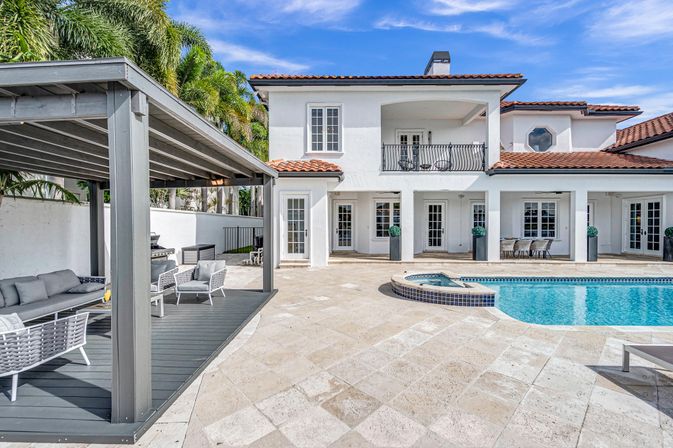 Sunny Mediterranean-style two-story villa with red tile roof, luxury backyard pool and raised spa, tiled patio, shaded grey pergola cabana lounge and palm trees