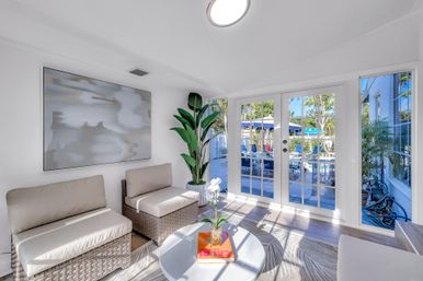 Bright, breezy poolside sunroom with wicker seating and neutral cushions, round white coffee table with an orchid and book, large abstract wall art, tall potted plant, and French doors opening to a sunny pool deck with lounge chairs and umbrellas.