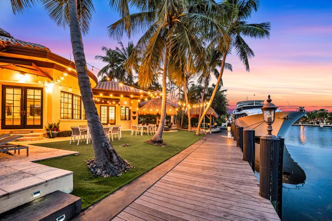 Tropical waterfront villa at sunset with string lights, palm trees, outdoor dining on a grassy patio and a docked yacht along a wooden boardwalk