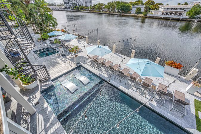Riverside modern pool deck with two-tier pool, submerged white lounge chairs, blue umbrellas, rows of sunbeds, string lights and spiral staircase overlooking a calm river