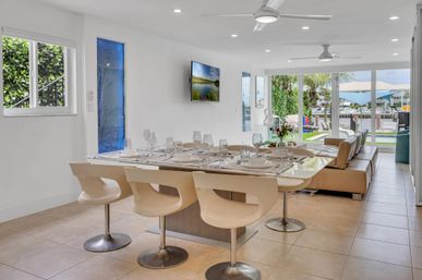 Bright modern open-plan dining and living room with island table set for six, sleek white swivel chairs, beige leather sofa, ceiling fans and large sliding glass doors opening to a sunlit backyard and waterfront dock.
