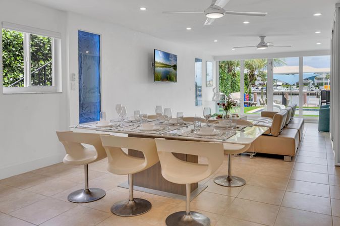 Bright modern open-plan dining and living room with island table set for six, sleek white swivel chairs, beige leather sofa, ceiling fans and large sliding glass doors opening to a sunlit backyard and waterfront dock.