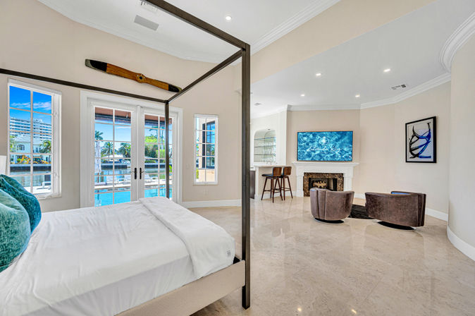 Waterfront master bedroom with four-poster bed, marble floors, glass doors to pool and canal, fireplace and cozy seating with wall-mounted TV and nautical propeller décor.