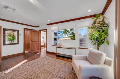 Sunlit coastal-style living room with hardwood floors and rich wood trim, open wooden double doors, neutral armchair, large potted fiddle-leaf fig, woven area rug, console table and wall-mounted TV showing a beach scene.