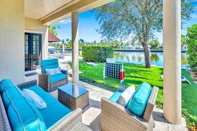 Covered waterfront patio with wicker seating and bright turquoise cushions, glass-top table, green lawn with oversized backyard games, palm trees and calm canal view under a sunny blue sky