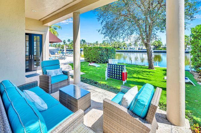 Covered waterfront patio with wicker seating and bright turquoise cushions, glass-top table, green lawn with oversized backyard games, palm trees and calm canal view under a sunny blue sky