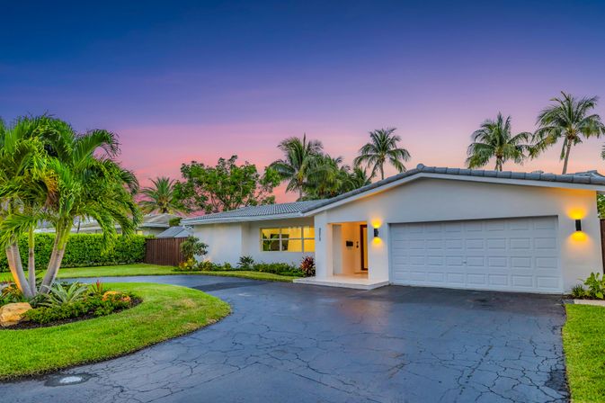 Twilight view of a white single-story suburban home with two-car garage, curved driveway, manicured lawn, tropical landscaping and palm trees against a pink‑purple sunset sky.