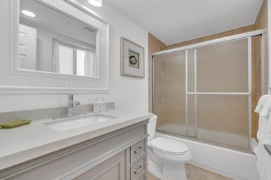 Bright modern home bathroom with white vanity and quartz countertop, chrome faucet and large mirror, toilet and bathtub with frosted sliding glass doors over beige tile, framed wall art and folded towels.