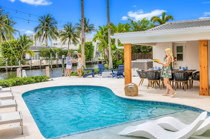 Tropical waterfront backyard with curved blue pool, in-pool loungers, palm trees and canal, covered patio dining and friends enjoying drinks