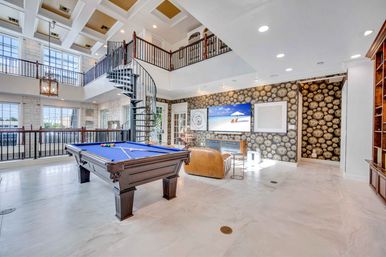 Sunlit two-story luxury game room with blue-felt pool table, spiral staircase to a mezzanine, large windows and coastal views