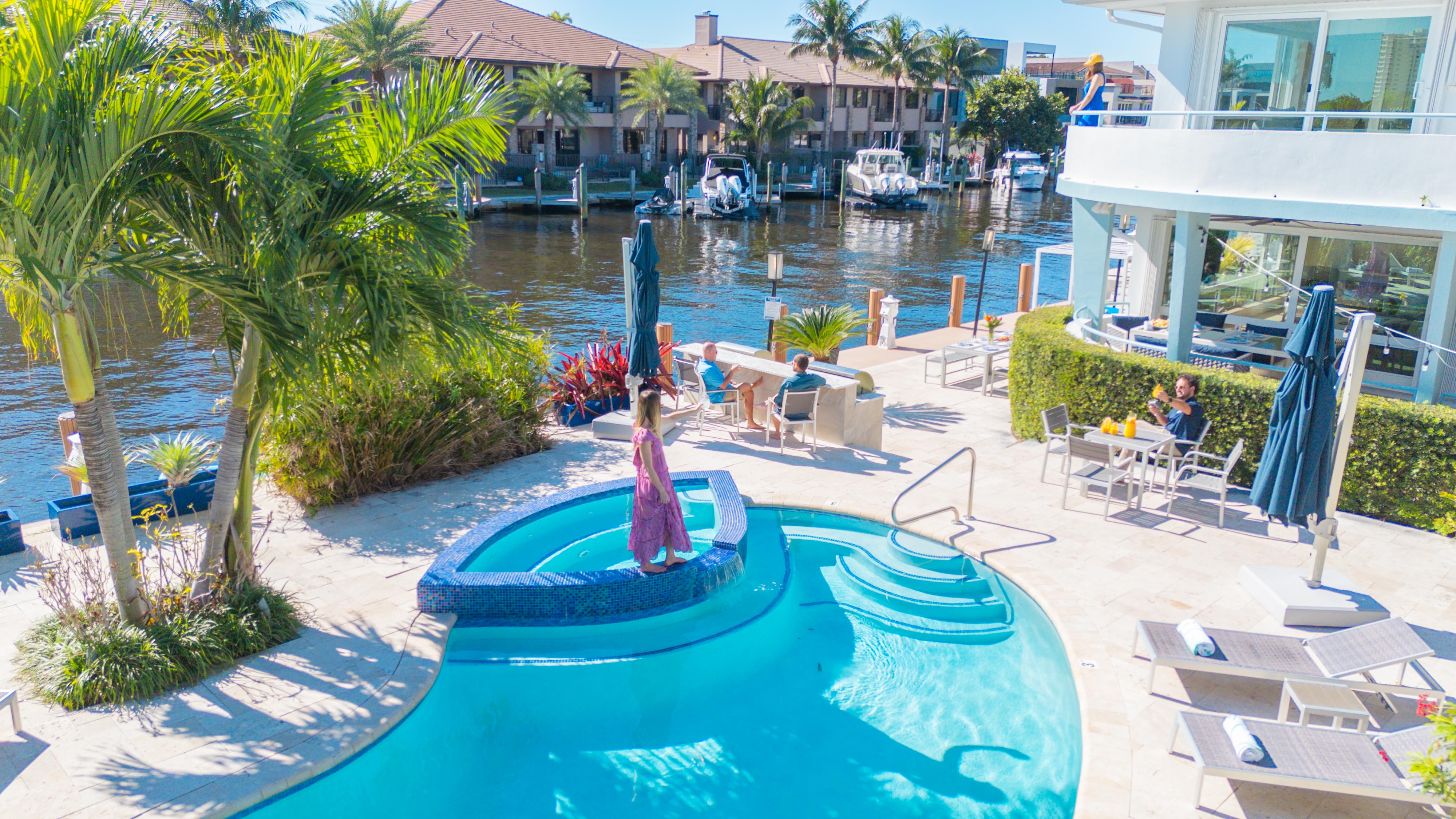 Sunny tropical canalfront pool with palm trees; woman in a pink dress on a mosaic spa ledge, docked boats and people dining on a waterfront patio.