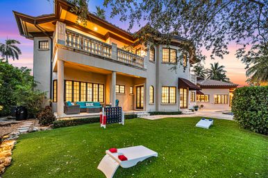 Sunset-lit Mediterranean-style two-story home with balcony and covered patio, glowing windows, teal outdoor seating, lush lawn with cornhole and giant Connect Four, palm trees