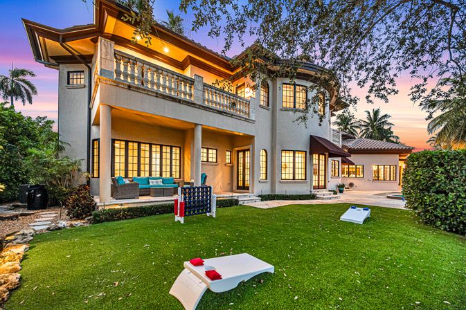 Sunset-lit Mediterranean-style two-story home with balcony and covered patio, glowing windows, teal outdoor seating, lush lawn with cornhole and giant Connect Four, palm trees