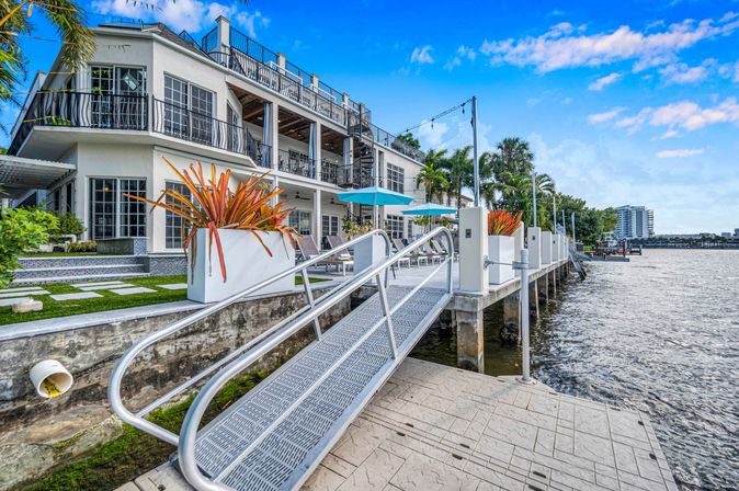 Sunny waterfront property: modern two-story white home with balconies, metal dock ramp to the river, tropical planters and blue patio umbrellas under a bright blue sky