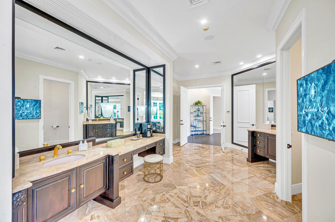 Spacious luxury master bathroom with polished marble floors, dark wood double vanities, gold fixtures, large mirrors, vanity stool and wall-mounted TV opening to a bright dressing area.