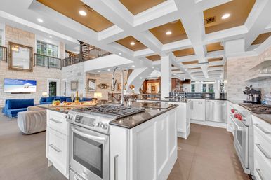 Spacious luxury open-concept kitchen with large center island, gas range and stainless appliances, coffered ceiling, stone accent walls and adjacent living area with blue sofas and a second-floor balcony.