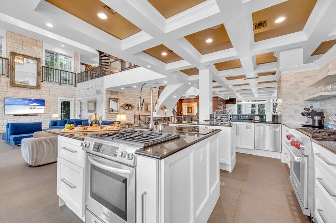 Spacious luxury open-concept kitchen with large center island, gas range and stainless appliances, coffered ceiling, stone accent walls and adjacent living area with blue sofas and a second-floor balcony.