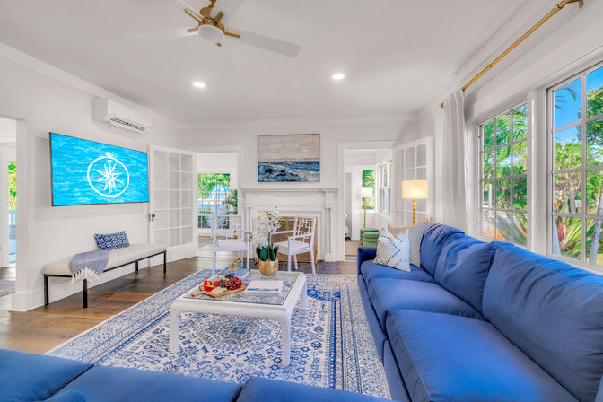Sunlit coastal-style living room with a deep-blue sectional, white fireplace, patterned blue-and-white rug, wall-mounted TV, French doors and large windows framed by tropical palms.