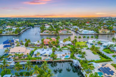 Aerial view of a South Florida waterfront neighborhood at sunset, showing palm-lined canals, luxury homes with private docks and boats, backyard pools and pastel skies.