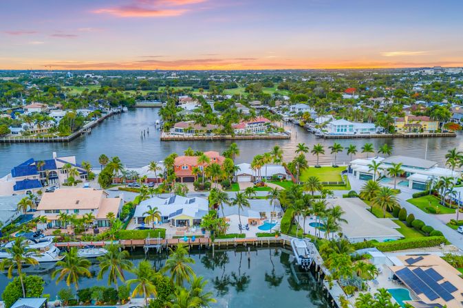 Aerial view of a South Florida waterfront neighborhood at sunset, showing palm-lined canals, luxury homes with private docks and boats, backyard pools and pastel skies.