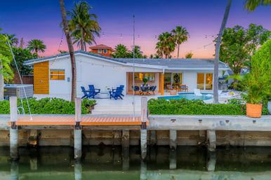 Tropical waterfront modern home at twilight with private dock, swimming pool, patio seating and string lights framed by palm trees and a purple-pink sunset sky