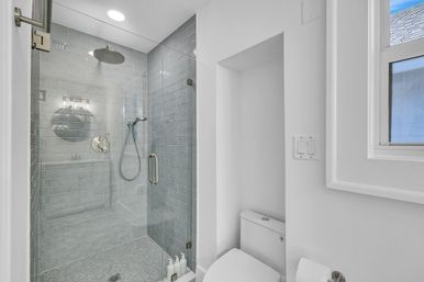 Bright modern bathroom with glass-enclosed gray subway-tiled shower, overhead rain shower and handheld, small hexagon mosaic floor, white toilet in a recessed nook and window letting in natural light.