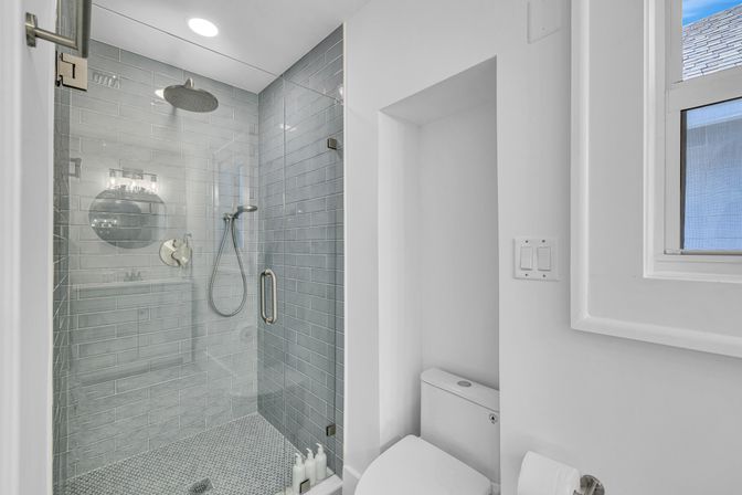 Bright modern bathroom with glass-enclosed gray subway-tiled shower, overhead rain shower and handheld, small hexagon mosaic floor, white toilet in a recessed nook and window letting in natural light.