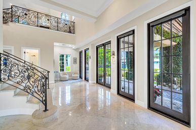 Sunlit luxury two-story foyer with ornate wrought-iron staircase, glossy marble floors and black-framed French doors opening to a leafy patio.