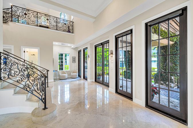 Sunlit luxury two-story foyer with ornate wrought-iron staircase, glossy marble floors and black-framed French doors opening to a leafy patio.