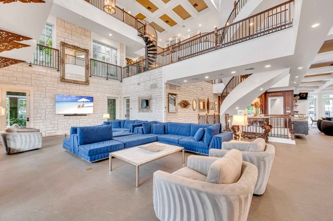 Sunlit luxury hotel lobby with a cobalt blue sectional and marble coffee table, plush armchairs, stone accent walls, mezzanine with ornate railings and a dramatic spiral staircase.
