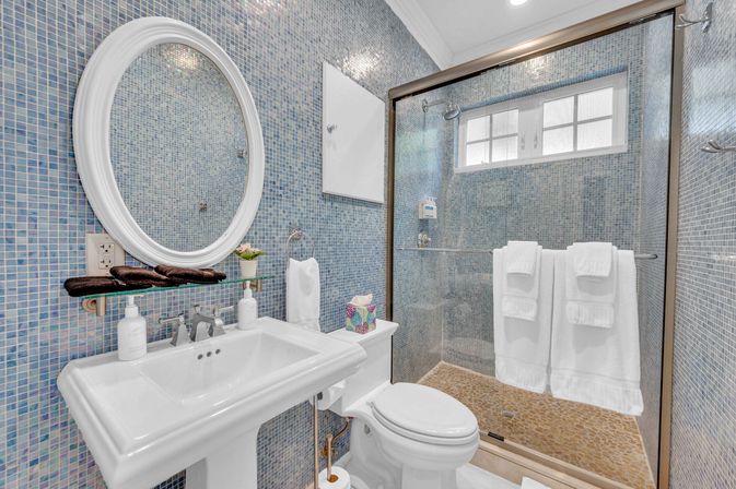 Bright blue mosaic bathroom with round white mirror, pedestal sink, toilet and glass-enclosed shower with pebble floor and neatly folded white towels