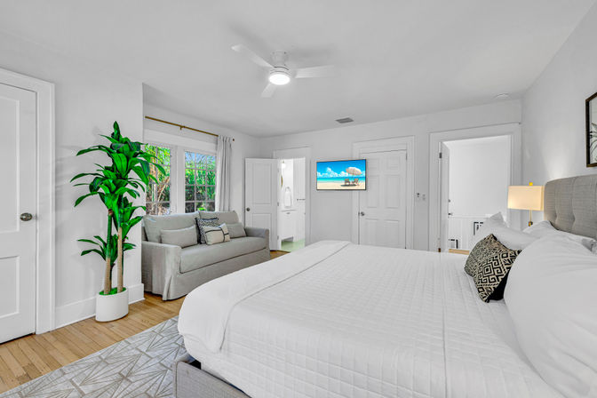 Bright coastal-inspired master bedroom in a modern home with king bed and white quilt, gray sofa by a garden-view window, tall potted plant, wall-mounted TV showing a beach scene, and en-suite bathroom.