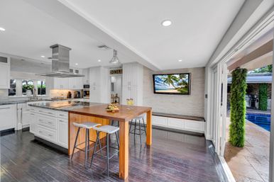 Open-concept modern white kitchen with stainless steel range hood, large wooden island table with stools, dark hardwood floors, wall-mounted TV and sliding doors opening to a pool patio with greenery.