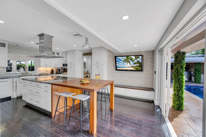 Open-concept modern white kitchen with stainless steel range hood, large wooden island table with stools, dark hardwood floors, wall-mounted TV and sliding doors opening to a pool patio with greenery.