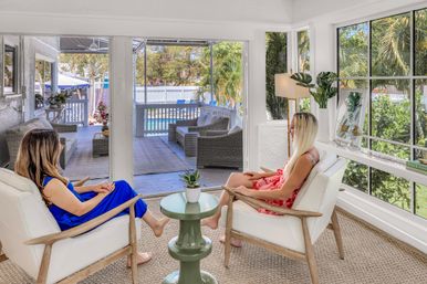 Two people in sundresses relax in a bright coastal sunroom, seated in white armchairs around a small green table, looking out through open doors to a palm-lined deck and swimming pool.