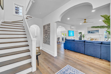 Bright open-concept entryway and living room with white wood staircase, arched doorways, wide-plank hardwood floors and a large navy sectional facing a fireplace and wall-mounted TV in a coastal-inspired home.